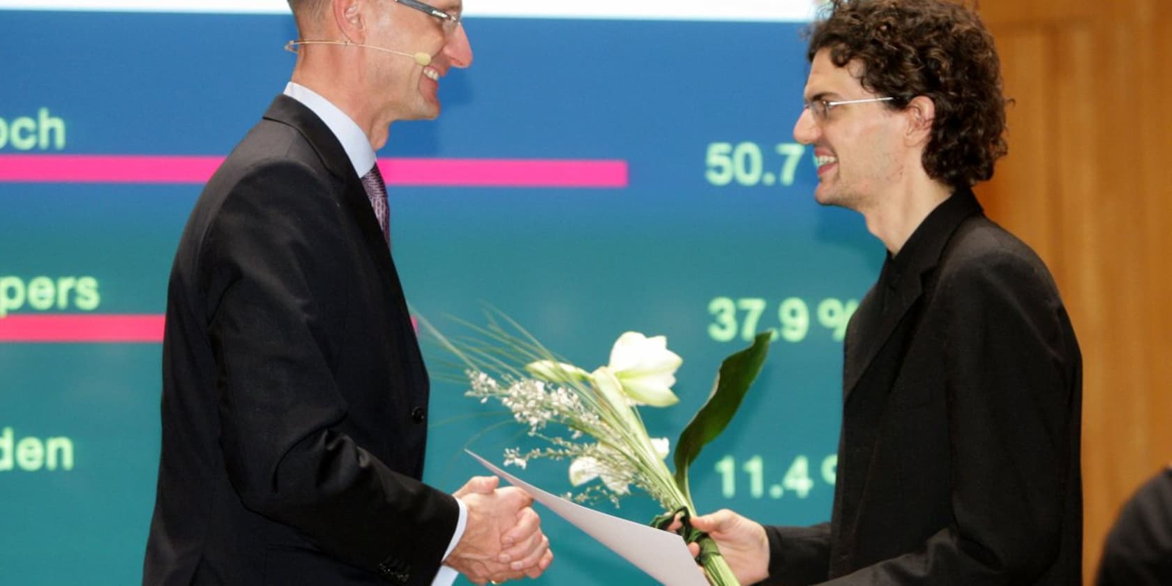 A man in a suit hands a certificate and flowers to another man in front of a screen displaying numbers.