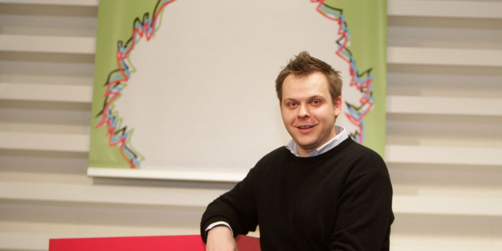 A man sits in front of a colorful poster and smiles at the camera.