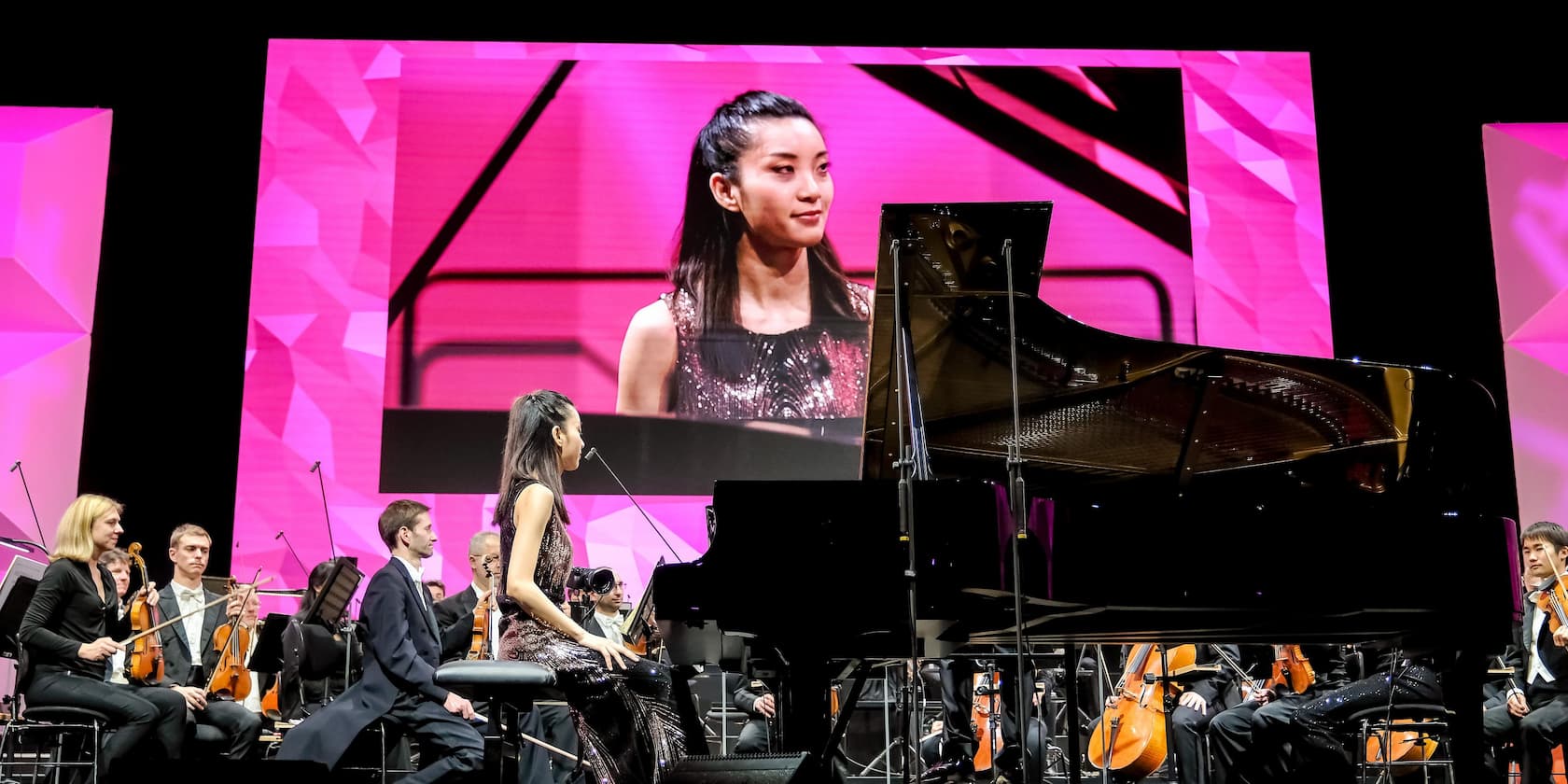 A woman plays the piano on stage, accompanied by an orchestra. She is shown on a large screen in the background.