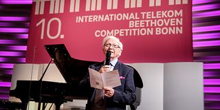 A man with white hair and glasses speaks into a microphone and holds a white leaflet. Behind him is an open grand piano.