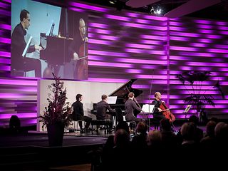 Three musicians in piano trio playing on stage with magenta background. The viewing angle is to the left of the stage.