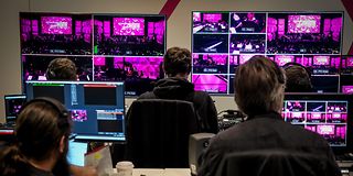 People sit in front of many screens that capture different angles of the stage in the hall. A lot of it is magenta.