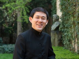 An Asian man in a black collared shirt smiles at the camera in a garden with a bust of Beethoven in the background.